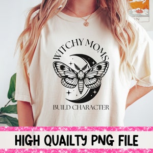 K&ouml;nnte beinhalten: Ein wei&szlig;es T-Shirt mit einem schwarzen Grafikdesign, das eine zunehmende Mondsichel mit einem Nachtfalter und Sternen zeigt. Der Text "Witchy Moms Build Character" ist unter dem Grafikdesign gedruckt.