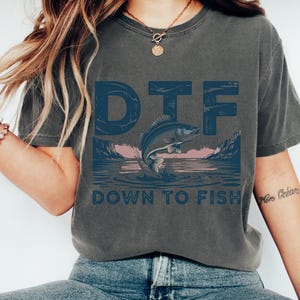 May include: Dark gray t-shirt with a blue graphic that says "DTF" above a jumping fish illustration. Below the fish, the text reads "DOWN TO FISH". The design features a lake scene with mountains and a pink sky.