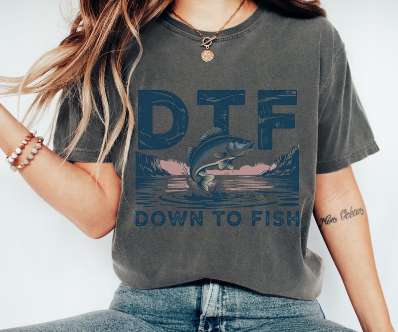 May include: Dark gray t-shirt with a blue graphic that says "DTF" above a jumping fish illustration. Below the fish, the text reads "DOWN TO FISH". The design features a lake scene with mountains and a pink sky.