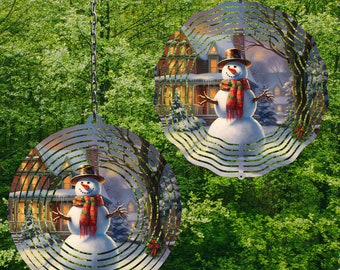 Könnte beinhalten: Zwei Windspiele aus Metall mit einer Winterszene, die einen Schneemann vor einem Haus mit beleuchteten Fenstern zeigt. Die Windspiele hängen an Ketten vor einem grünen, belaubten Hintergrund.