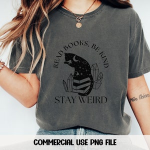May include: A dark gray t-shirt with a black graphic of a cat sitting on a stack of books. The graphic also includes crystals, stars, and a crescent moon. The text "Read Books, Be Kind, Stay Weird" is printed on the shirt.