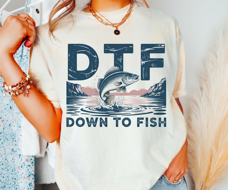 May include: Cream-colored t-shirt with a graphic featuring a fish leaping from water, mountains, and the text "DTF DOWN TO FISH" in navy blue. The design is centered on the shirt.
