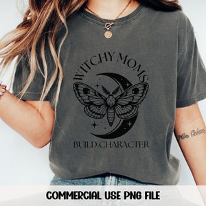 K&ouml;nnte beinhalten: Ein dunkelgraues T-Shirt mit einem schwarzen Grafikdesign, das eine Sichelmond, einen Nachtfalter und Sterne zeigt. Der Text "Witchy Moms Build Character" ist auf das Shirt gedruckt.