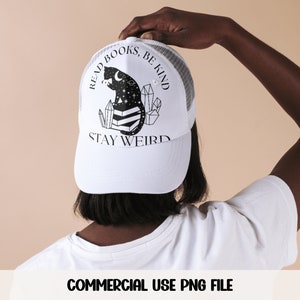 May include: White trucker hat with a black and white illustration of a cat sitting on books with crystals and stars. The text "Read Books, Be Kind, Stay Weird" is printed on the hat.