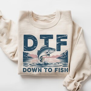 May include: A cream-colored sweatshirt with a graphic design featuring a jumping fish and the text "DTF DOWN TO FISH" in navy blue. The design includes a lake scene with mountains and water ripples.