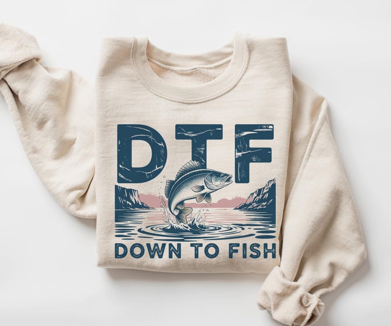 May include: A cream-colored sweatshirt with a graphic design featuring a jumping fish and the text "DTF DOWN TO FISH" in navy blue. The design includes a lake scene with mountains and water ripples.