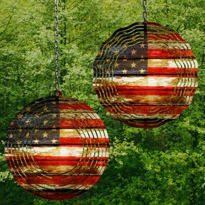 May include: Two red, white, and blue American flag wind spinners hanging from chains against a green leafy background.