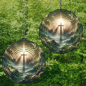 May include: Two metal wind chimes with a cross and clouds design. The chimes are hanging from chains against a green leafy background.