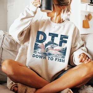 May include: Cream-colored sweatshirt with the text "DTF DOWN TO FISH" in blue. The graphic features a fish jumping out of the water with a mountain and water scene. The sweatshirt is a relaxed fit.