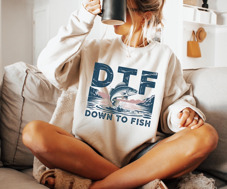 May include: Cream-colored sweatshirt with the text "DTF DOWN TO FISH" in blue. The graphic features a fish jumping out of the water with a mountain and water scene. The sweatshirt is a relaxed fit.
