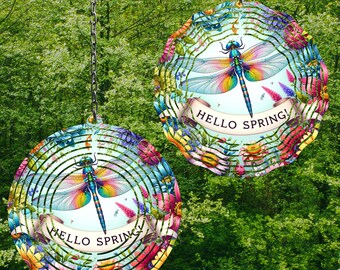May include: Two colorful metal wind spinners with a dragonfly design and the text "Hello Spring!" on a white banner. The spinners are hanging from chains against a green leafy background.