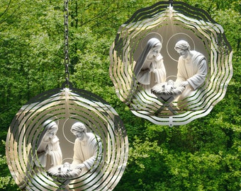 May include: Two metal wind spinners with a circular design featuring a white nativity scene. The spinners are hanging from chains against a green leafy background.