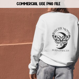 K&ouml;nnte beinhalten: Wei&szlig;er Pullover mit einem schwarz-wei&szlig;en Motiv eines Nachtfalters und eines Halbmondes. Der Schriftzug lautet "Witchy Moms Build Character".