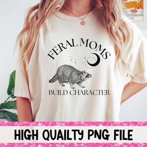 Pode incluir: Uma camiseta branca com o texto "Feral Moms Build Character" em preto. Uma ilustração em preto e branco de um guaxinim com uma lua crescente e estrelas acima está representada na camiseta.