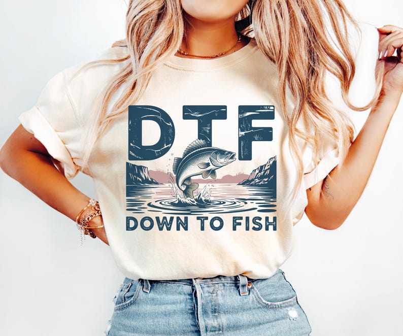 May include: Cream-colored t-shirt featuring a blue graphic with a jumping fish, water, and mountains. The text "DTF DOWN TO FISH" is printed below the graphic in a distressed font. The shirt is a casual style.