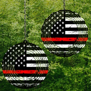 Puede incluir: Dos carillones redondos de metal con un diseño de bandera estadounidense roja, blanca y azul. La bandera está estilizada para parecerse a un anillo de árbol. Los carillones cuelgan de cadenas contra un fondo de follaje verde.