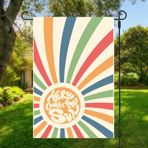 Può includere: Una bandiera da giardino con un design a raggi di sole retrò in tonalità di arancione, rosso, giallo, verde e blu. I raggi di sole circondano un cerchio bianco con il testo "Here Comes the Sun" in un font retrò.