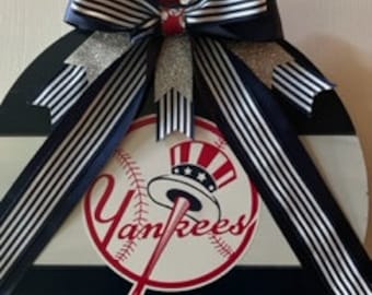 May include: A round wooden door hanger with a navy blue, white, and silver striped bow. The center of the door hanger features a white and red New York Yankees logo with the words "Yankees" in red.