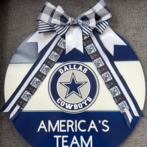 Puede incluir: Un letrero redondo de madera azul, blanco y gris con el logotipo de los Dallas Cowboys y el texto "Dallas Cowboys" y "America's Team". El letrero est&aacute; decorado con una cinta a cuadros en blanco y negro.