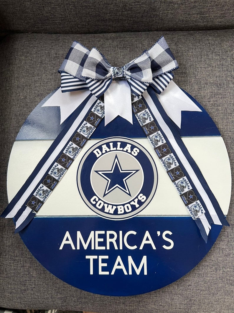 Puede incluir: Un letrero redondo de madera azul, blanco y gris con el logotipo de los Dallas Cowboys y el texto "Dallas Cowboys" y "America's Team". El letrero est&aacute; decorado con una cinta a cuadros en blanco y negro.
