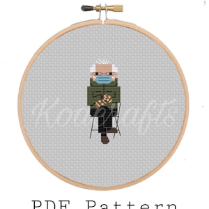 May include: Cross stitch pattern of a person wearing a face mask and sitting on a chair. The person is wearing a green jacket and black pants. The pattern is in a circular hoop.