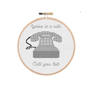 May include: A black and white cross-stitch embroidery hoop with the text "You're in a cult. Call your dad." and a black and white cross-stitch image of a rotary phone.