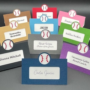 May include: A set of 10 baseball-themed place cards with different names and titles printed on them. The place cards are made of textured cardstock in various colors, each featuring a baseball graphic with red stitching.