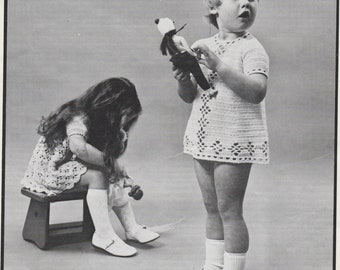May include: Two children in white crochet dresses. One holds a doll, the other sits on a small wooden stool. The dresses feature short raglan sleeves and a diamond pattern around the lower edge and front.