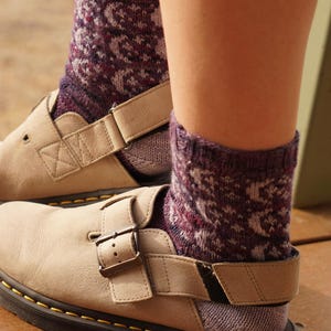 May include: A pair of beige leather slip-on shoes with a buckle strap across the top and a thick black sole with yellow stitching, worn with purple and white patterned knit socks that feature a repeating geometric design around the ankles.