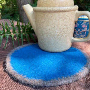 May include: A beige ceramic teapot with a lid sits on a round, blue felt coaster with a gray border. The teapot has a rounded body, a curved spout, and a handle. The coaster is on a brown surface.