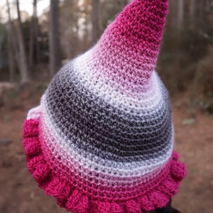 May include: A handmade crochet hat with a pointed top and a ruffled brim. The hat features a gradient of colors, transitioning from hot pink to white and gray. The hat is a unique accessory.
