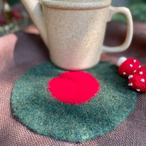 May include: A beige ceramic teapot with a matching lid sits atop a green and red felt trivet. The trivet has a circular design with a red center. A small, red mushroom decoration with white spots is also visible.