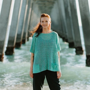 Puede incluir: Una mujer con un top de crochet turquesa y pantalones negros se encuentra de pie bajo un muelle con el océano detrás de ella.