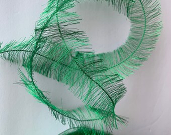 May include: A spool of green, sparkly, fringed ribbon. The ribbon is curled into a circle and the spool is labeled "Our Arts".