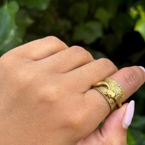 May include: A gold-coloured ring with a snake design. The ring features a detailed floral pattern and a snake head. The ring is worn on a finger, showcasing its intricate design and craftsmanship. The ring is a piece of jewellery.