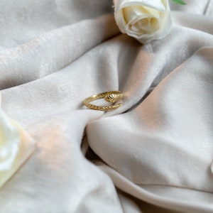 May include: A gold snake ring with textured detailing. The ring is coiled, with the snake's head at the top. It is set against a cream-coloured, silky fabric, with a white rose in the background.