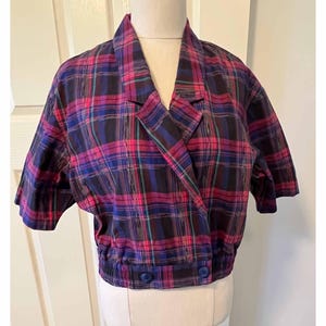 May include: A cropped plaid jacket in shades of purple, pink, and blue. The jacket has a notched collar, short sleeves, and two blue buttons. The fabric features a classic plaid pattern.