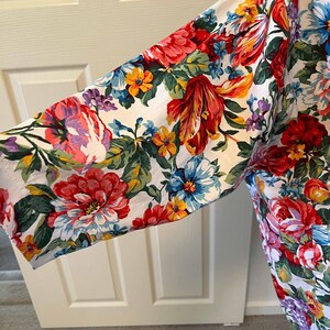 May include: A floral print garment featuring a vibrant array of red, pink, yellow, and blue flowers on a white background. The sleeve is visible, showcasing the colorful botanical pattern. The fabric appears to be lightweight.