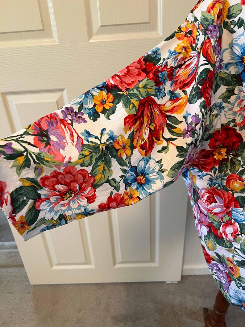 May include: A floral print garment featuring a vibrant array of red, pink, yellow, and blue flowers on a white background. The sleeve is visible, showcasing the colorful botanical pattern. The fabric appears to be lightweight.