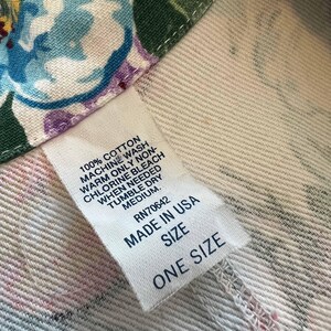 May include: Close-up of a white fabric with a floral pattern in blue, purple, and pink. A white tag reads "100% Cotton" and "Made in USA". The tag also indicates care instructions and "One Size".
