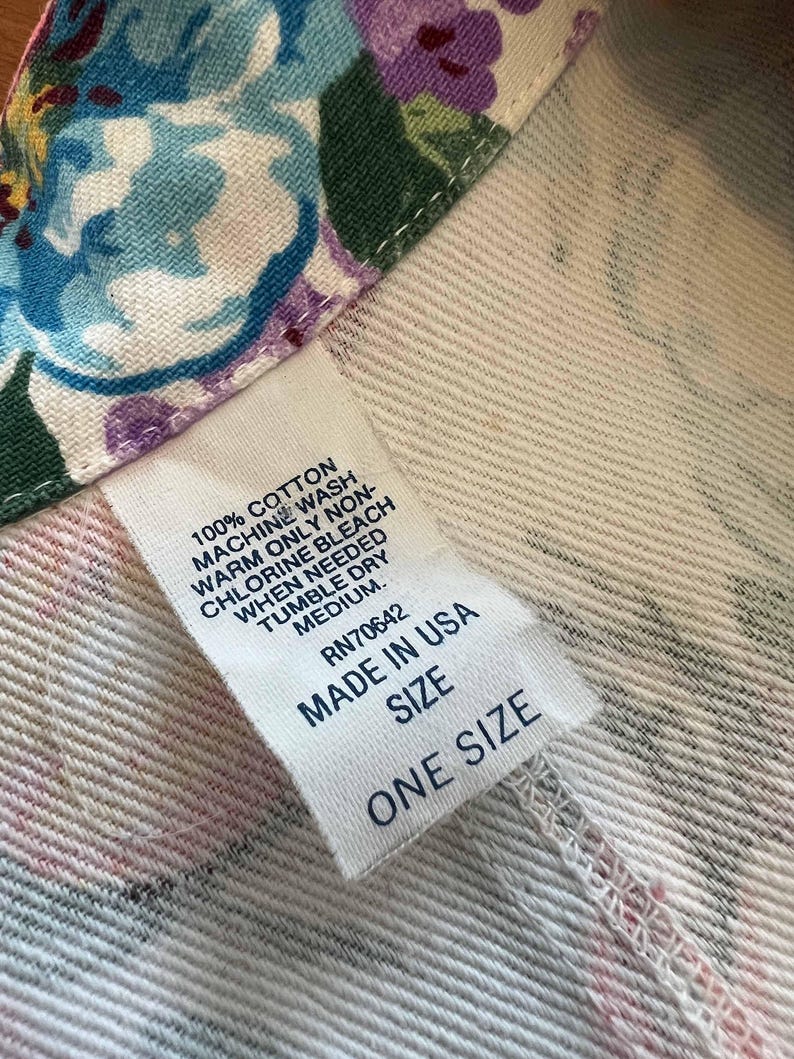 May include: Close-up of a white fabric with a floral pattern in blue, purple, and pink. A white tag reads "100% Cotton" and "Made in USA". The tag also indicates care instructions and "One Size".