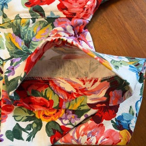 May include: Close-up of a garment with a vibrant floral pattern. The fabric features large, colorful roses in shades of red, pink, yellow, and blue against a white background. The garment's seams and folded edges are visible.