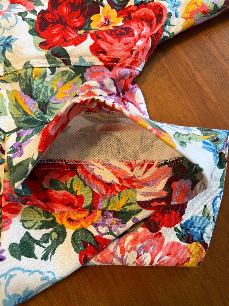 May include: Close-up of a garment with a vibrant floral pattern. The fabric features large, colorful roses in shades of red, pink, yellow, and blue against a white background. The garment's seams and folded edges are visible.