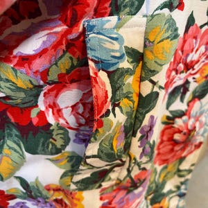May include: Close-up of a floral fabric with a pocket detail. The fabric features a vibrant pattern of red, pink, yellow, and purple flowers with green leaves on a white background. The pocket is outlined with light blue stitching.