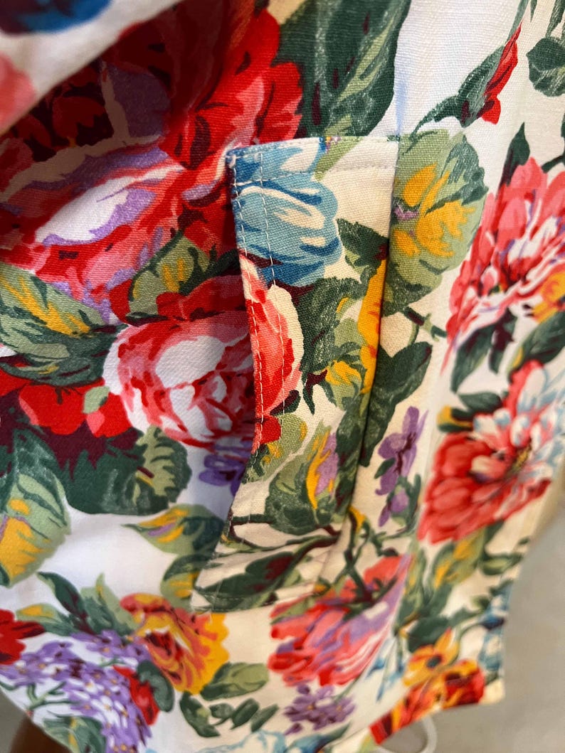 May include: Close-up of a floral fabric with a pocket detail. The fabric features a vibrant pattern of red, pink, yellow, and purple flowers with green leaves on a white background. The pocket is outlined with light blue stitching.