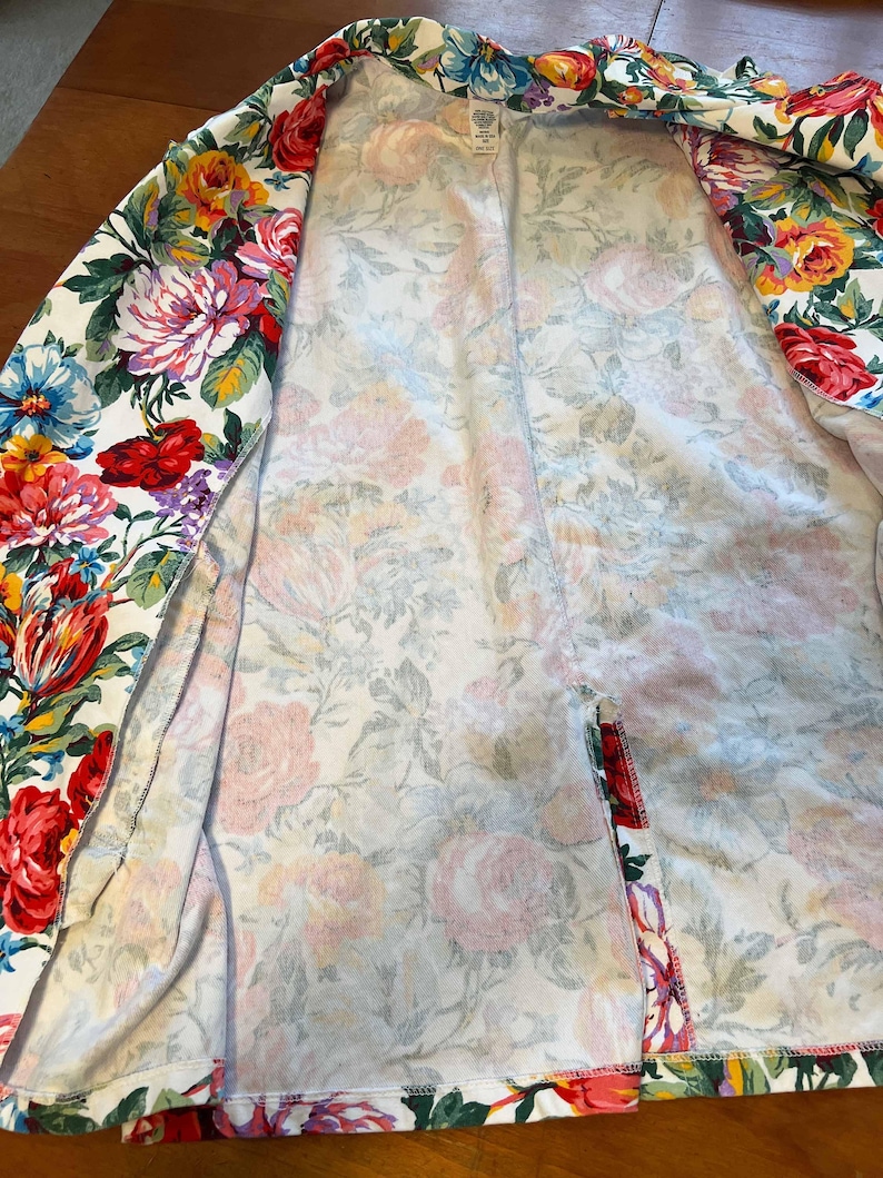 May include: A white floral print blazer featuring a vibrant array of red, pink, yellow, and blue flowers. The open blazer showcases a soft, lightweight fabric with a colorful botanical design, perfect for adding a touch of spring to any outfit.