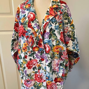 May include: A floral print blazer with a white base, adorned with a colourful pattern of red, pink, yellow, blue, and purple flowers. The blazer has a lapel collar, long sleeves, and a single-button closure. The garment is displayed on a mannequin.