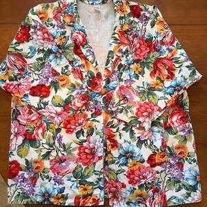 May include: A floral print blazer featuring a vibrant mix of red, pink, blue, yellow, and purple flowers on a white background. The blazer has a classic collar, button closure, and short sleeves, perfect for a spring or summer look.