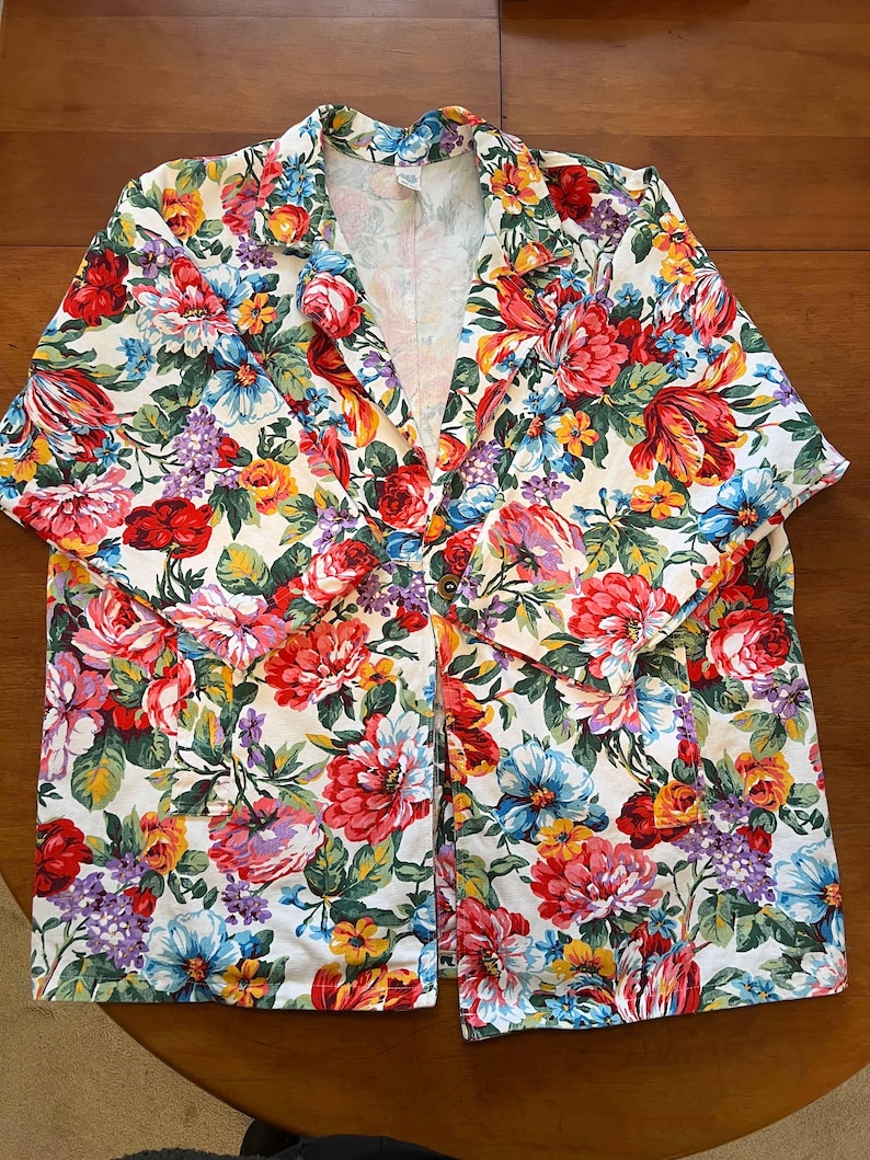 May include: A floral print blazer featuring a vibrant mix of red, pink, blue, yellow, and purple flowers on a white background. The blazer has a classic collar, button closure, and short sleeves, perfect for a spring or summer look.