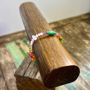 May include: A multi-coloured gemstone bracelet featuring pink, green, yellow, and orange beads. The bracelet is presented on a wooden stand, highlighting its vibrant colours and unique design. The bracelet is approximately 18 cm in length.
